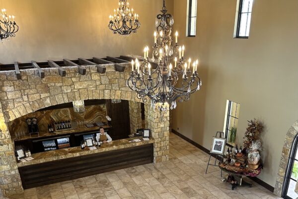 A spacious, elegant lobby with stone walls, high ceilings, large chandeliers, and a warmly lit reception desk where a staff member is standing. Decor includes artwork, wine bottles, and a festive autumn display.