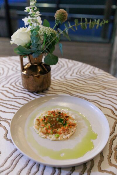 A round plate with a finely plated dish featuring a small mound of diced vegetables and possibly fish, garnished with herbs and surrounded by a ring of green sauce. A vase with white flowers sits in the background on a patterned tablecloth.