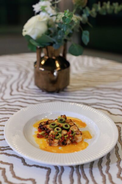 A plated gourmet dish with sliced olives, vegetables, and sauce, presented on a white plate. A decorative vase with white flowers sits blurred in the background on a patterned tablecloth.