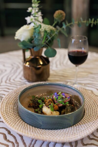 A plated gourmet meal with various small portions, garnished with herbs, sits on a woven placemat beside a glass of red wine and a small vase of white flowers on a patterned tablecloth.