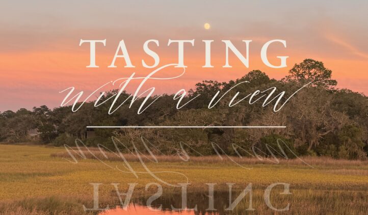A sunset sky in shades of pink and orange reflects on a calm marsh with tall grasses; text over the image reads, Tasting with a view.