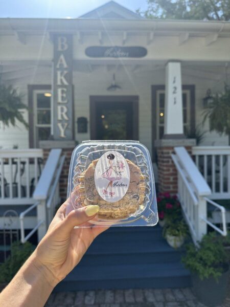 A hand holds a clear plastic container of cookies with a stylish woman illustration in front of a white bakery building with a BAKERY sign and blue steps leading to the entrance.