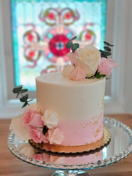 A white and pink ombre cake decorated with fresh roses and eucalyptus leaves sits on a glass cake stand, with a colorful stained glass window in the background.