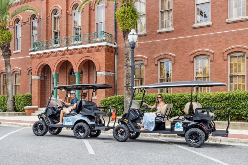 Two people sit in separate black golf carts parked on a street in front of a historic red brick building with arched windows and palm trees. Both drivers are smiling and wearing sunglasses.
