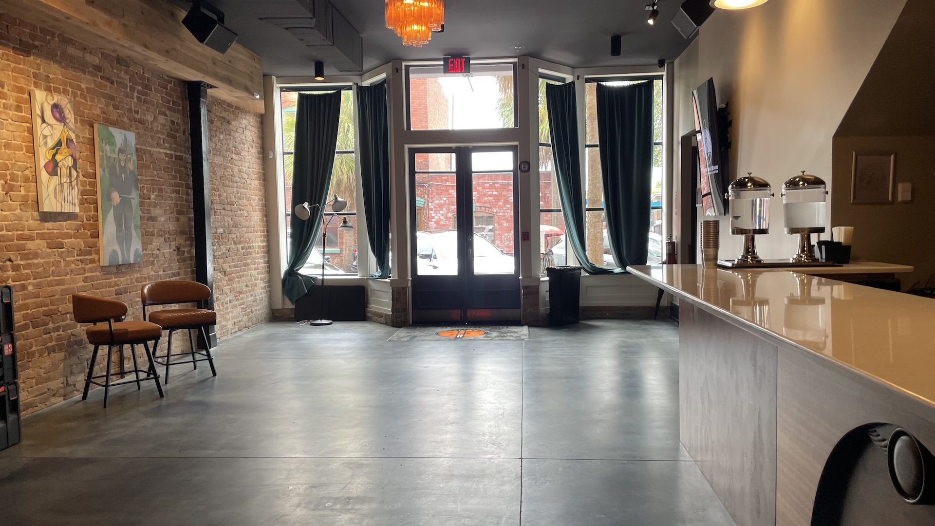 A modern, empty cafe with exposed brick walls, tall windows with dark curtains, two brown chairs, art on the walls, and a counter with two drink dispensers. Natural light fills the space.