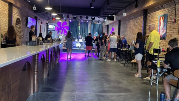 People gather inside a modern bar with exposed brick walls and colorful lighting, facing a stage with musical instruments. Some are standing, others sit at tables; artwork decorates the walls.