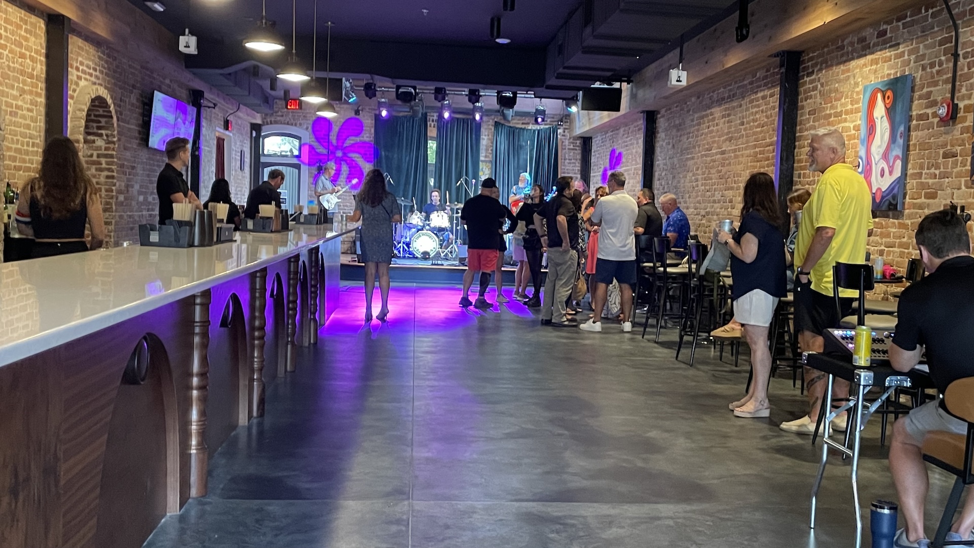 People gather inside a modern bar with exposed brick walls and colorful lighting, facing a stage with musical instruments. Some are standing, others sit at tables; artwork decorates the walls.