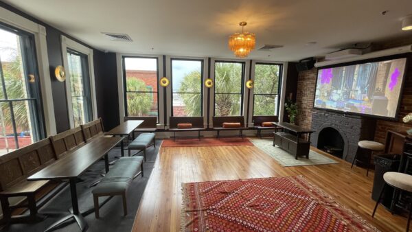 A cozy, modern lounge with large windows, wooden floors, benches and chairs along the walls, a chandelier, a brick fireplace, and a projector screen displaying a colorful scene. Palm trees are visible outside.