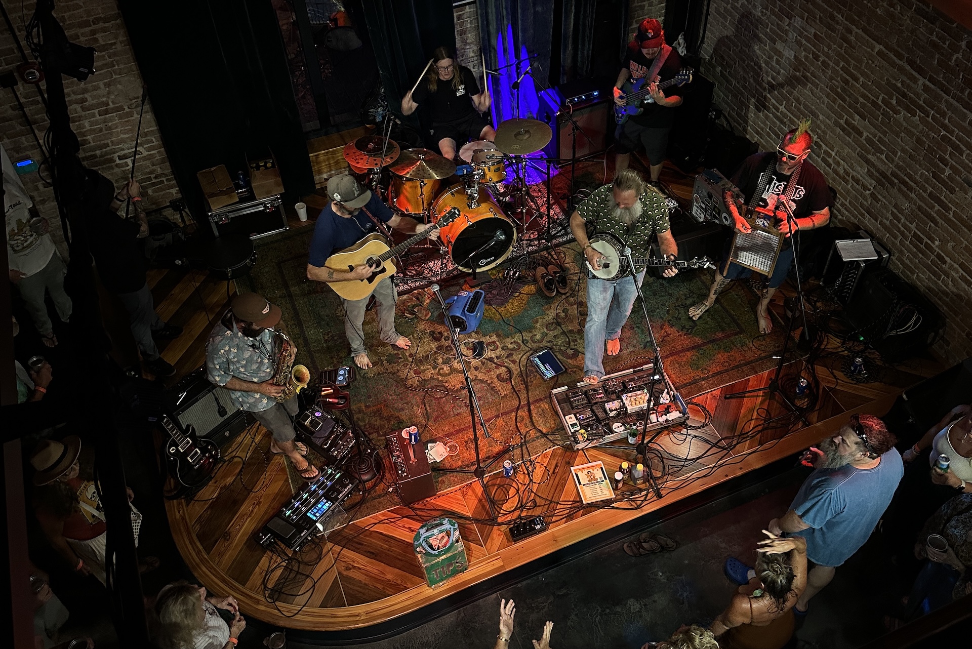 A band performs on a small stage with guitars, banjo, drums, and a washboard, surrounded by cables and equipment, while an enthusiastic audience watches from below. The scene is set in a cozy, brick-walled venue.