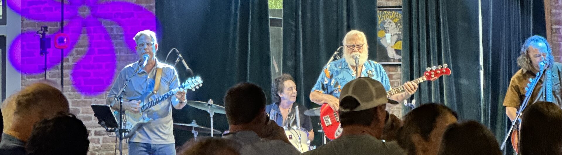 A band performs on stage in front of a crowd, with blue and purple lights shining. The musicians play guitars and drums, and a pink flower design is projected on a brick wall behind them. Audience members watch and enjoy the music.