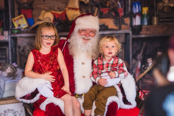 Two young children, one in a red dress and the other in a plaid shirt, sit on Santa Claus’s lap. Santa is smiling, dressed in his classic red suit, in front of a festive, rustic holiday backdrop.
