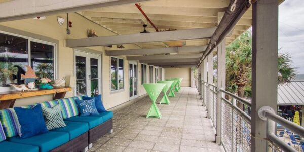 A covered outdoor patio with green cocktail tables, blue cushioned benches with decorative pillows, and a view of palm trees and rooftops below. The space is bright and overlooks an outdoor area.