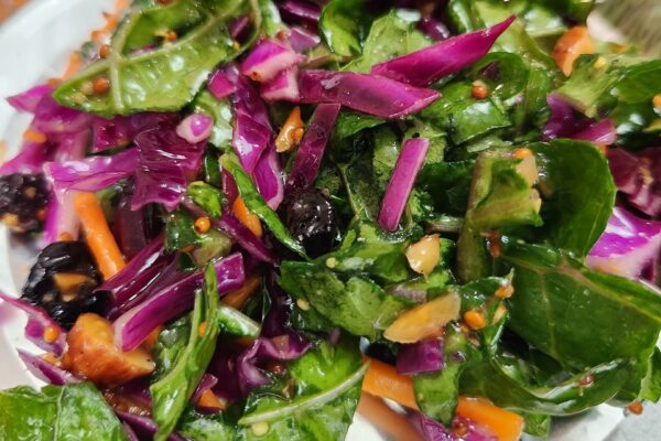 Close-up of a colorful salad with leafy greens, purple cabbage, carrot strips, black olives, and visible mustard seeds, all mixed with a vinaigrette dressing.