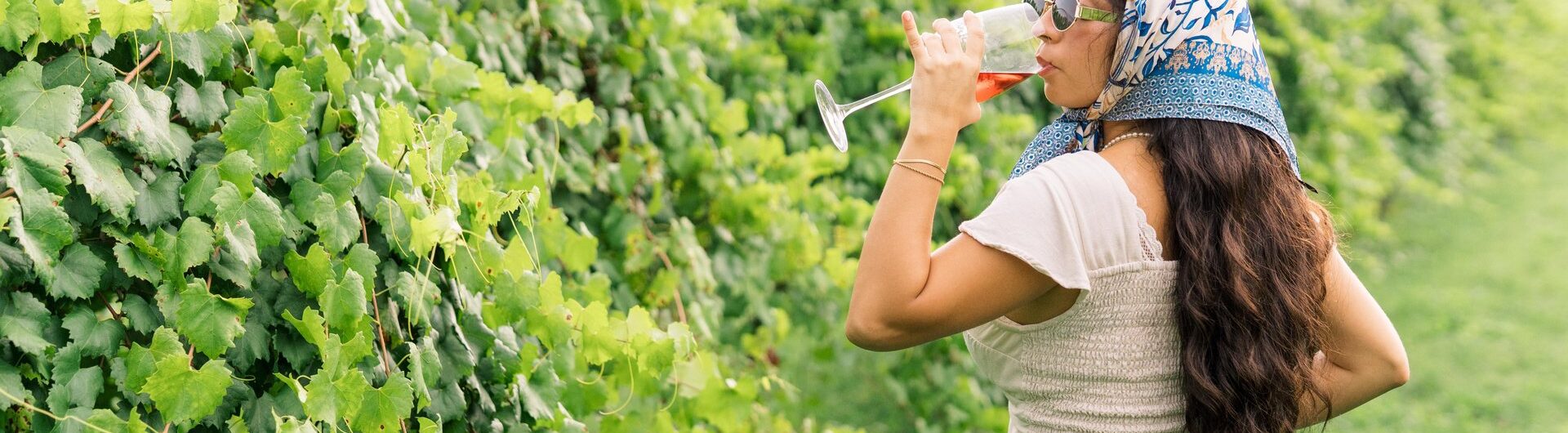 A woman wearing a headscarf, sunglasses, and jeans stands in a vineyard, drinking from a wine glass. Lush green grapevines line her side, and she faces away from the camera on a bright, sunny day.