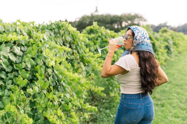 A woman wearing a headscarf, sunglasses, and jeans stands in a vineyard, drinking from a wine glass. Lush green grapevines line her side, and she faces away from the camera on a bright, sunny day.
