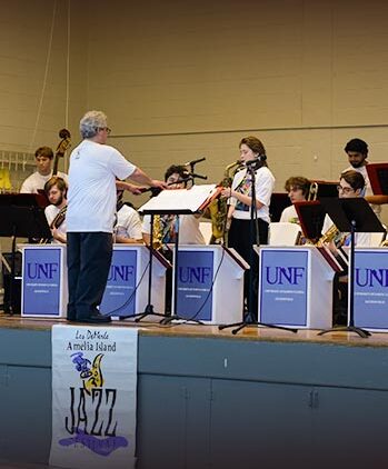 A jazz band of young musicians performs on stage with a conductor leading them. They play various instruments including saxophones and brass. The stage has UNF signs and a banner reading “Jazz Festival.”.