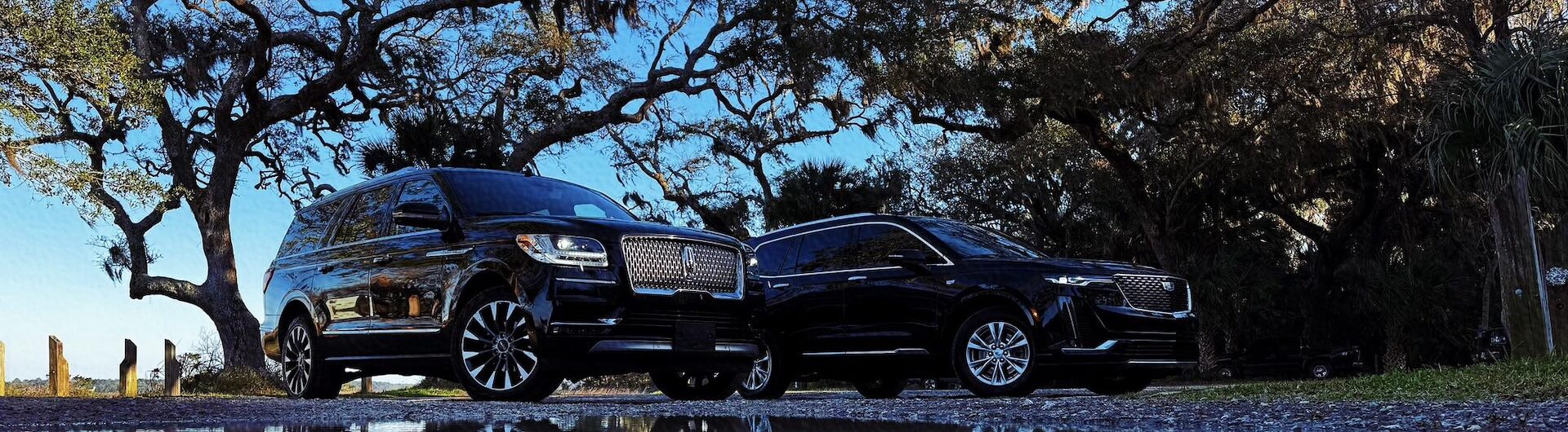 Two luxury black SUVs parked under oak trees on Amelia Island with reflections in a puddle beneath a clear blue sky