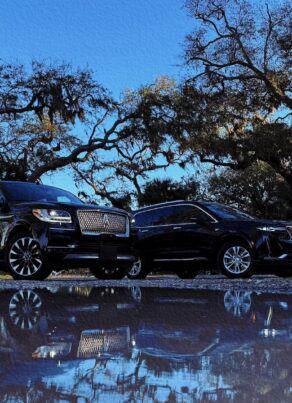 Two luxury black SUVs parked under oak trees on Amelia Island with reflections in a puddle beneath a clear blue sky