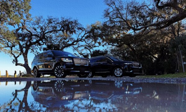 Two luxury black SUVs parked under oak trees on Amelia Island with reflections in a puddle beneath a clear blue sky