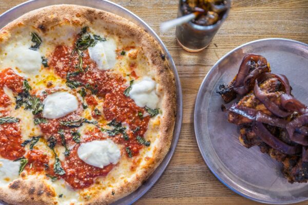 A Margherita pizza with tomato sauce, mozzarella, and basil sits on a metal tray next to a plate of grilled meat topped with red onions and a glass of cola on a wooden table.