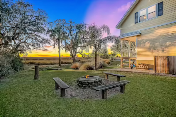 A cozy backyard scene at sunset with a stone fire pit surrounded by three wooden benches. Tall grasses, palm trees, and a yellow house with a porch are in the background. The sky glows with vibrant blue, pink, and orange hues.