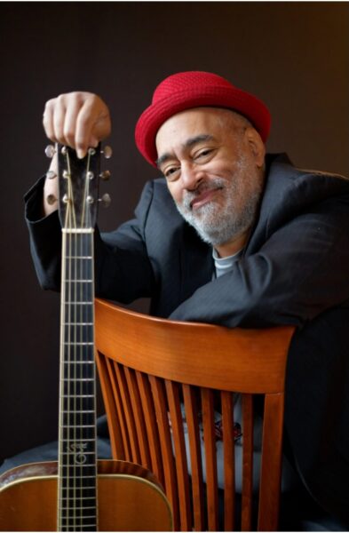 A smiling man with a gray beard, wearing a red hat and dark blazer, sits backward on a wooden chair, resting one arm on top of an acoustic guitar in front of him, against a dark background.