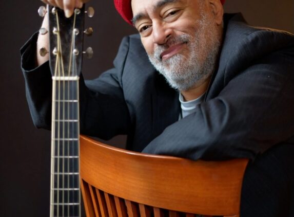A smiling man with a gray beard, wearing a red hat and dark blazer, sits backward on a wooden chair, resting one arm on top of an acoustic guitar in front of him, against a dark background.