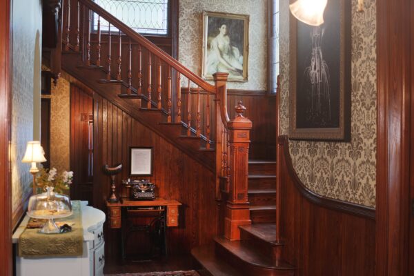 A vintage interior with a wooden staircase, ornate wallpaper, a framed portrait, and a small desk with an old typewriter and gramophone. A lamp and glass cake stand sit on a white side table.