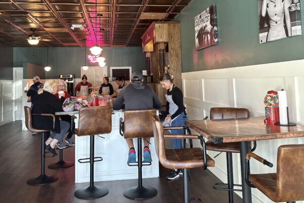 A retro-style diner with a few people sitting at the counter and tables. The decor features brown leather chairs, gumball machines, photos on the walls, and a green and white color scheme with a copper ceiling.
