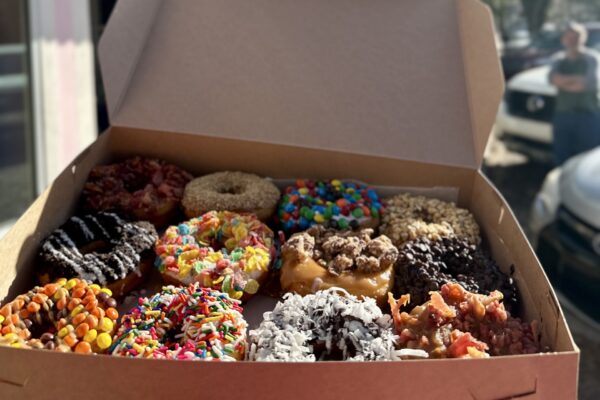 A box of twelve assorted donuts, each topped with colorful toppings like sprinkles, candy pieces, chocolate chips, coconut flakes, and bacon bits, sits open in sunlight.