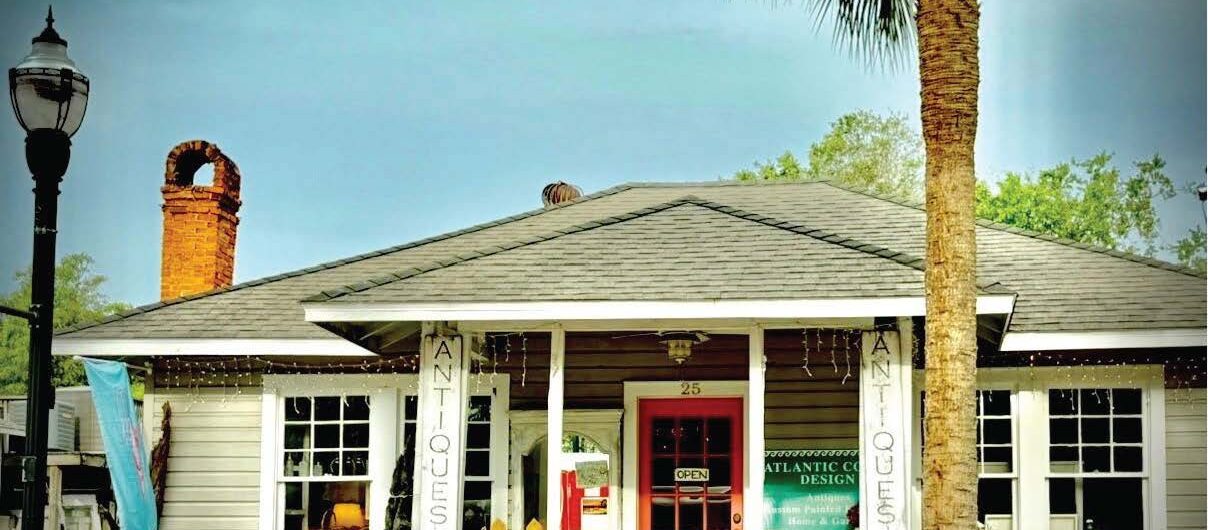 A small antique shop with a pink door, surrounded by plants and benches, sits under a palm tree. ANTIQUES signs hang by the entrance, and a lamppost is on the left. The scene has a cozy, welcoming feel.