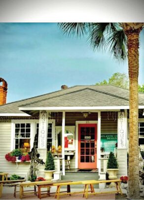 A small antique shop with a pink door, surrounded by plants and benches, sits under a palm tree. ANTIQUES signs hang by the entrance, and a lamppost is on the left. The scene has a cozy, welcoming feel.