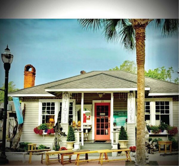 A small antique shop with a pink door, surrounded by plants and benches, sits under a palm tree. ANTIQUES signs hang by the entrance, and a lamppost is on the left. The scene has a cozy, welcoming feel.