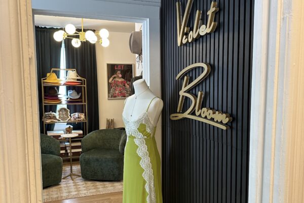 A mannequin in a green dress with lace trim stands by a black wall with gold Violet Bloom signage, near a cozy room with green chairs, a rug, shelves of hats, and a framed poster on the wall.
