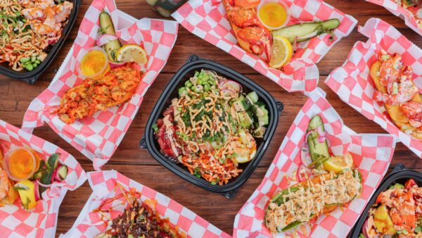 Several trays of colorful, fresh seafood dishes and poke bowls are arranged on a wooden table with red and white checkered paper, featuring ingredients like fish, avocado, veggies, sauces, and lemon slices.