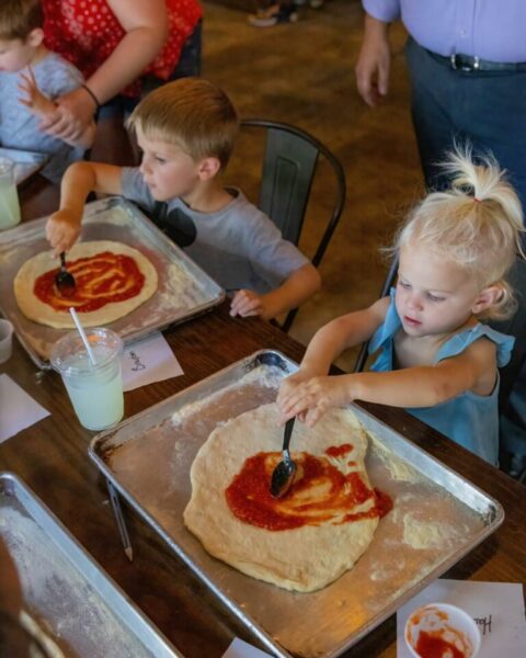 Two young children spread tomato sauce on pizza dough with spoons at a table. Each has a tray with dough and a drink nearby. Other people stand and sit around them, creating a busy, casual atmosphere.