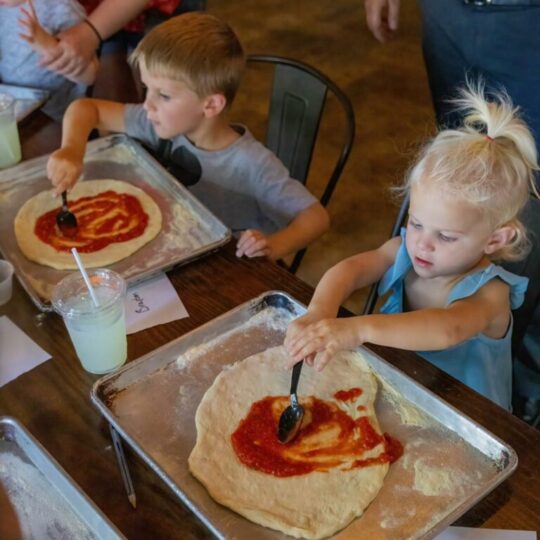 Two young children spread tomato sauce on pizza dough with spoons at a table. Each has a tray with dough and a drink nearby. Other people stand and sit around them, creating a busy, casual atmosphere.