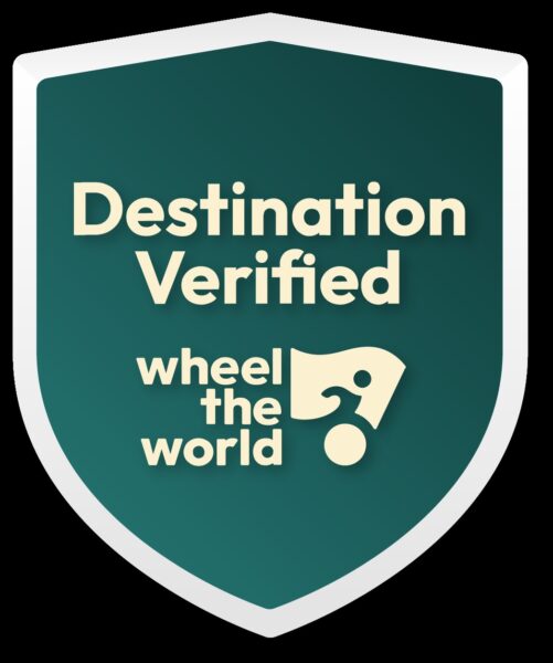 A green shield-shaped badge with the text Destination Verified above the logo and words wheel the world. The logo includes a stylized figure in a wheelchair.