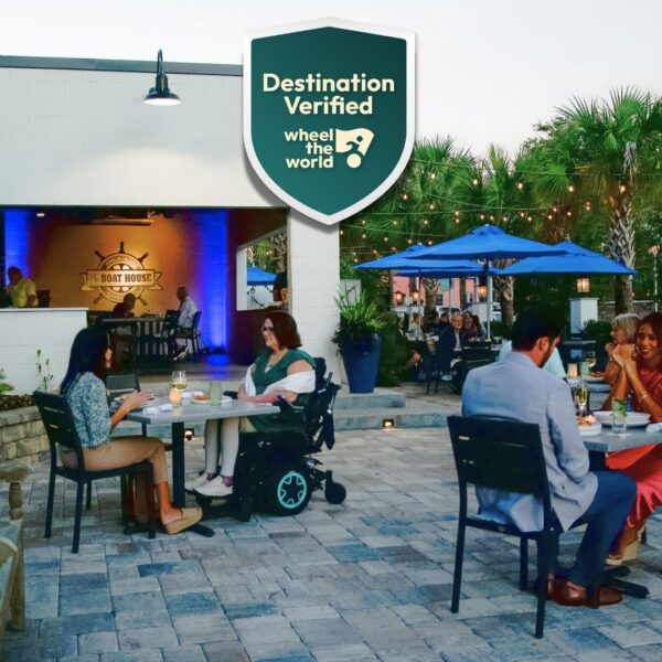 People dining outdoors at a restaurant patio with string lights and palm trees. A woman using a wheelchair is at one table. A Destination Verified Wheel the World accessibility badge is shown at the top.