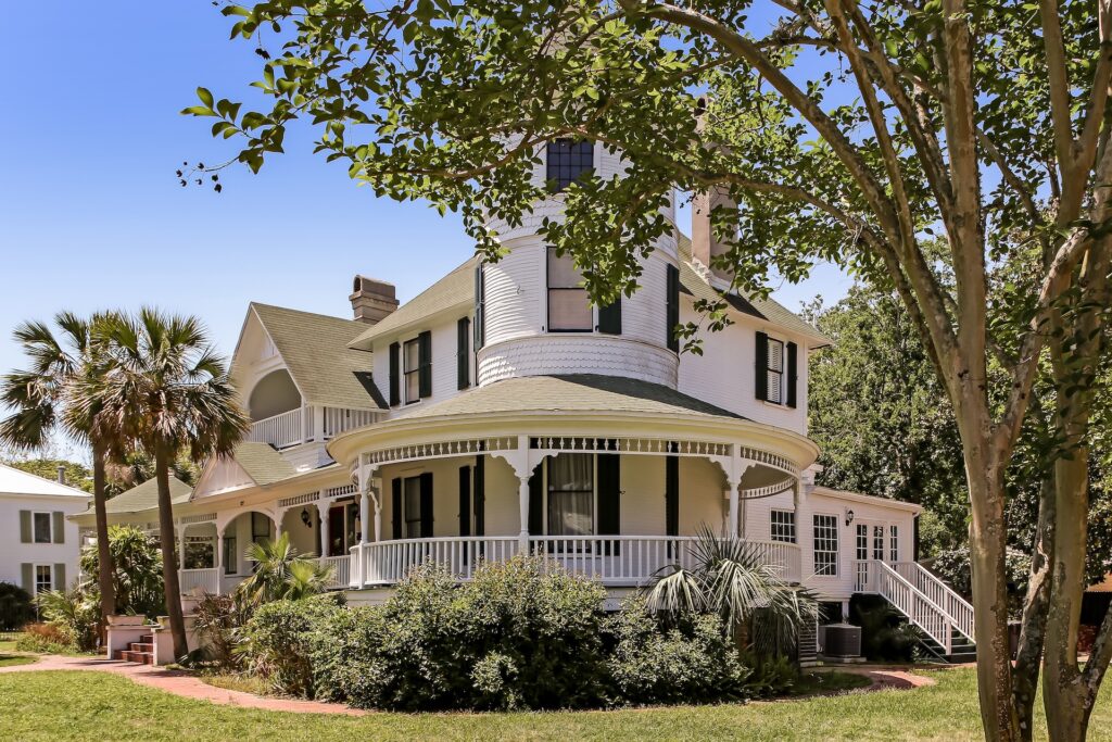 A large white Victorian-style house with a wraparound porch, green roof, and a round turret, surrounded by lush greenery, palm trees, and a clear blue sky in the background.