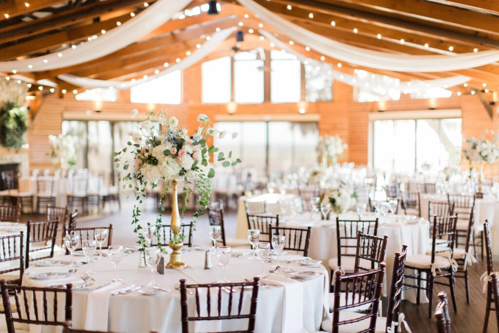 Elegant wedding reception hall with round tables covered in white linens, floral centerpieces, wooden chairs, and draped fabric and string lights hanging from a wooden beamed ceiling, creating a warm, festive atmosphere.