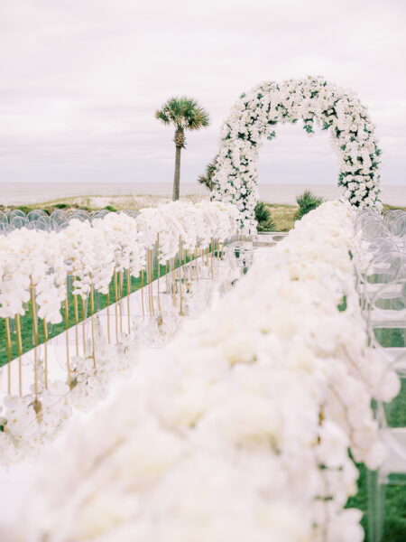 A wedding aisle lined with white flowers leads to a floral arch outdoors near a beach, with a single palm tree in the background under a cloudy sky.