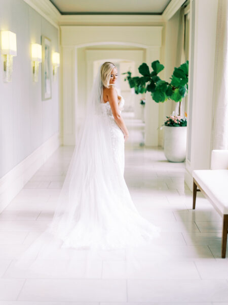 A bride in a white wedding dress and long veil stands in a bright hallway, looking over her shoulder. Sunlight streams in from large windows, highlighting her dress and creating a soft, elegant atmosphere.