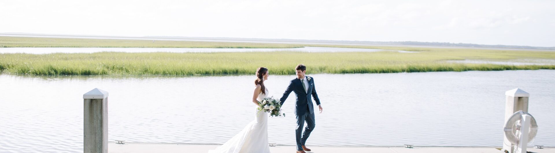 A bride and groom walk hand-in-hand on a dock over calm water, with grassy marshland in the background and bright sunlight illuminating the scene.