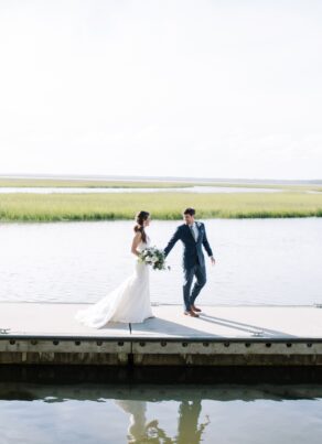 A bride and groom walk hand-in-hand on a dock over calm water, with grassy marshland in the background and bright sunlight illuminating the scene.