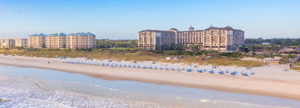 A wide sandy beach with rows of white chairs and umbrellas, gentle waves, and several large beachfront hotels and buildings surrounded by greenery under a clear blue sky.