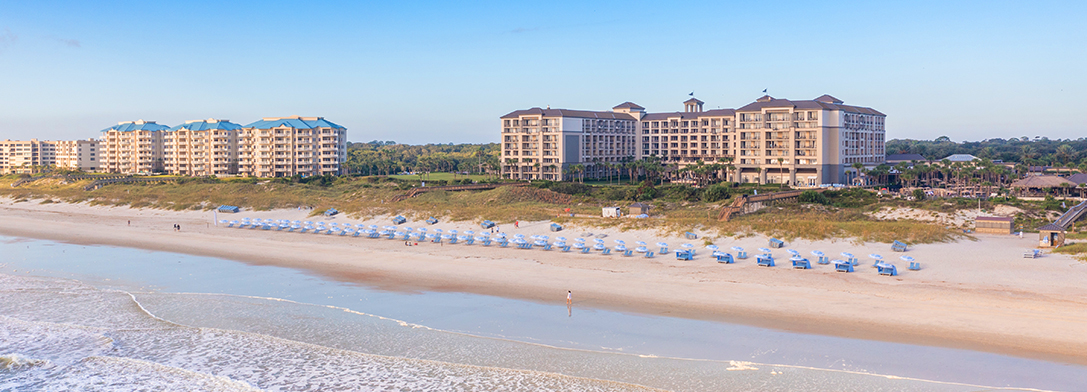 A wide sandy beach with rows of white chairs and umbrellas, gentle waves, and several large beachfront hotels and buildings surrounded by greenery under a clear blue sky.