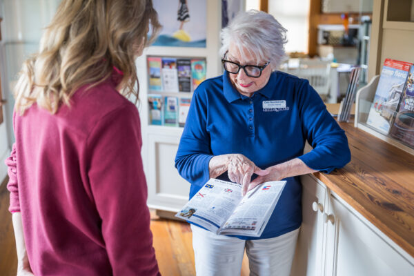 An older woman with white hair and glasses, wearing a blue shirt and name tag, shows a magazine to a younger woman in a red top in a cozy indoor setting.