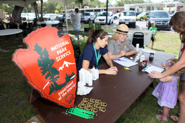 Two women sit at a table with a National Park Service sign, talking to a child and adult. The table has brochures, pencils, and badges. People and cars are visible in the background at an outdoor event.
