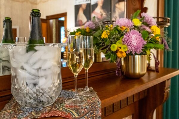 A bottle of champagne in an ice bucket and two filled champagne flutes sit on a patterned cloth atop a wooden mantle, next to a gold vase holding a colorful bouquet of flowers.